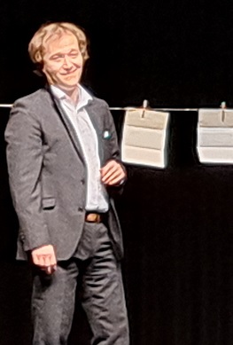 Schauspieler im Anzug auf Theaterbühne neben aufgehängten Blättern.