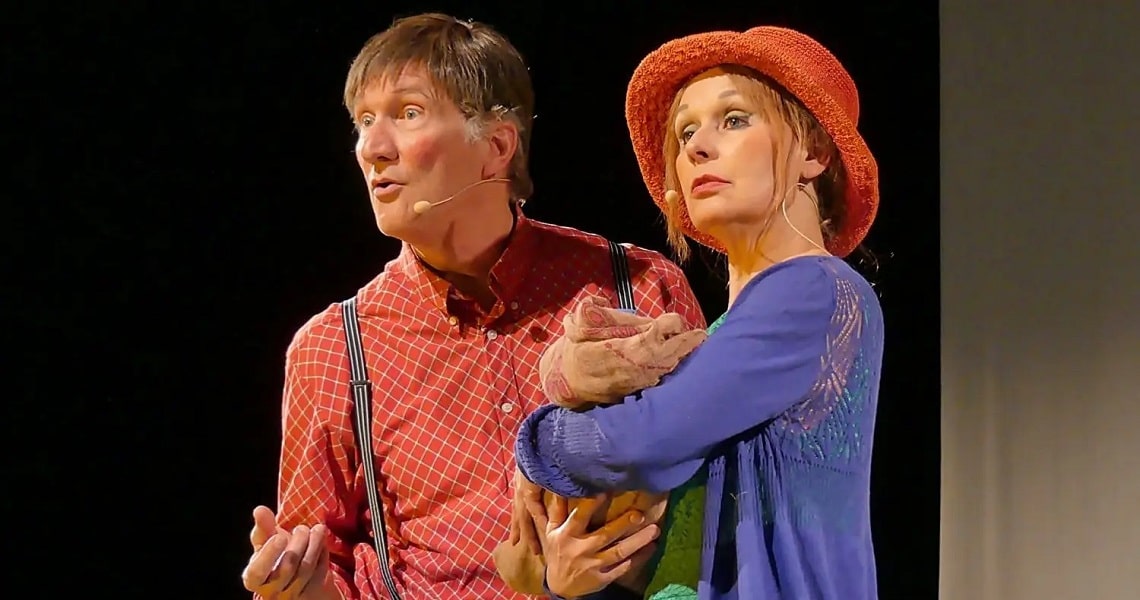 Stefan Österle gefällt als Sebastian Blau Zwei Schauspieler auf der Bühne mit Mikrofonen, er in rotem Hemd, sie mit orangefarbenem Hut und blauem Kleid, halten gemeinsam eine Stoffpuppe.