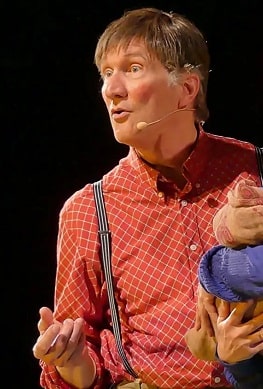 Schauspieler auf der Bühne in rotem Hemd und Hosenträgern, mit Mikrofon und ausdrucksstarker Geste während einer Theateraufführung.