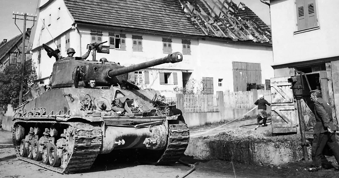 Panzer fährt durch das Dorf Rosswälden, während im Hintergrund ein Haus brennt und Rauch aufsteigt – historische Kriegsaufnahme aus dem Zweiten Weltkrieg.