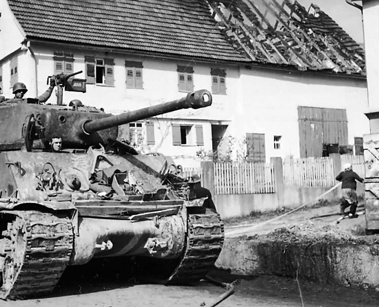 Panzer fährt durch das Dorf Rosswälden, während im Hintergrund ein Haus brennt und Rauch aufsteigt – historische Kriegsaufnahme aus dem Zweiten Weltkrieg.