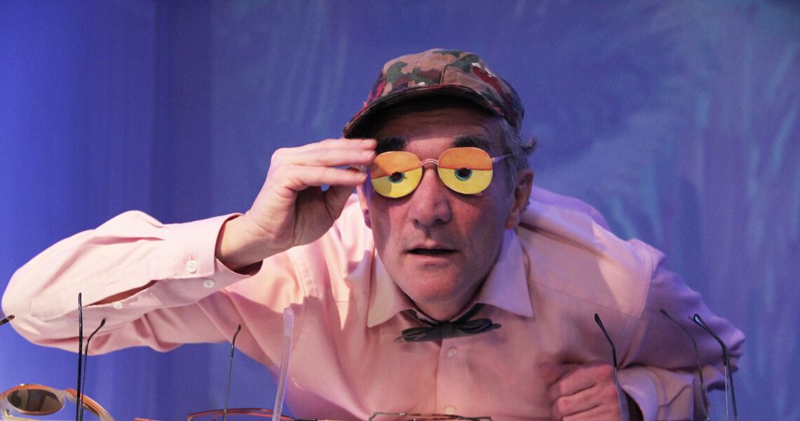 Ein Schauspieler hebt eine bunte Brille mit orange-gelben Gläsern und blickt neugierig ins Publikum. Szene aus dem Nachmittagstheater in der Kulturhalle Süßen.
