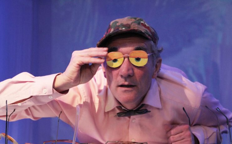 Ein Schauspieler hebt eine bunte Brille mit orange-gelben Gläsern und blickt neugierig ins Publikum. Szene aus dem Nachmittagstheater in der Kulturhalle Süßen.