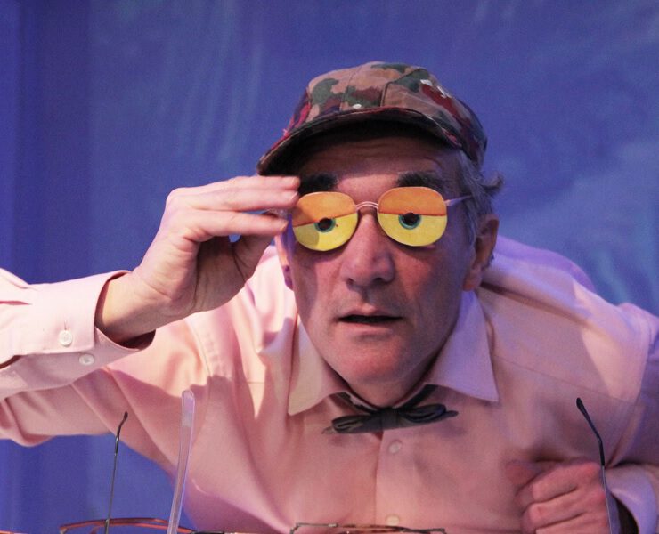 Ein Schauspieler hebt eine bunte Brille mit orange-gelben Gläsern und blickt neugierig ins Publikum. Szene aus dem Nachmittagstheater in der Kulturhalle Süßen.