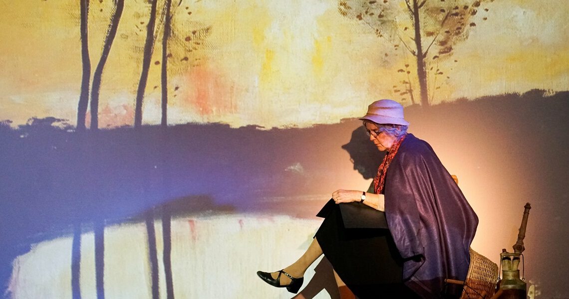 Eine ältere Frau sitzt vor einer poetischen, herbstlich projizierten Kulisse und liest nachdenklich. Aufführung des Nachmittagstheaters in der Kulturhalle Süßen.