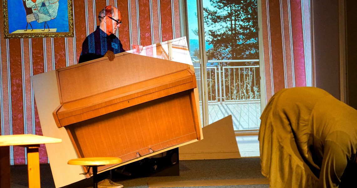 Ein Mann trägt ein übergroßes, kartoniertes Klavier auf der Bühne, daneben eine in Stoff gehüllte Figur. Szene aus dem Nachmittagstheater in der Kulturhalle Süßen mit surrealem Humor.