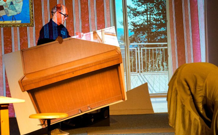 Ein Mann trägt ein übergroßes, kartoniertes Klavier auf der Bühne, daneben eine in Stoff gehüllte Figur. Szene aus dem Nachmittagstheater in der Kulturhalle Süßen mit surrealem Humor.