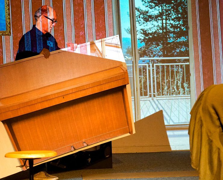 Ein Mann trägt ein übergroßes, kartoniertes Klavier auf der Bühne, daneben eine in Stoff gehüllte Figur. Szene aus dem Nachmittagstheater in der Kulturhalle Süßen mit surrealem Humor.