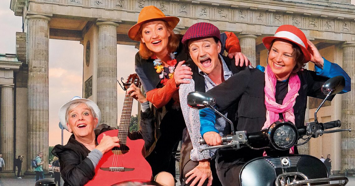 Vier fröhliche Darsteller:innen mit Hüten, Gitarre und Motorroller vor dem Brandenburger Tor. Heitere Szene aus dem Nachmittagstheater in der Kulturhalle Süßen.