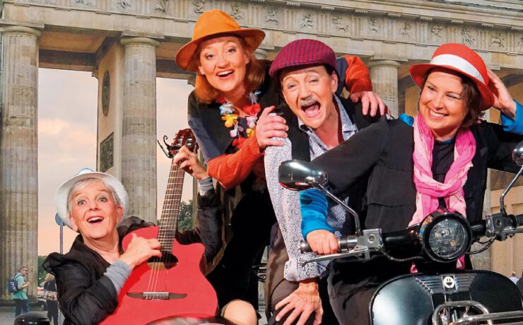 Vier fröhliche Darsteller:innen mit Hüten, Gitarre und Motorroller vor dem Brandenburger Tor. Heitere Szene aus dem Nachmittagstheater in der Kulturhalle Süßen.