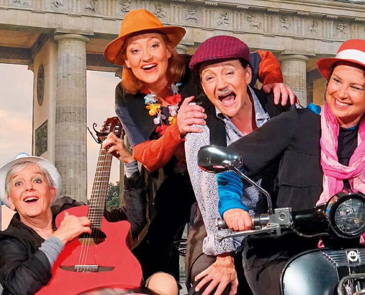 Vier fröhliche Darsteller:innen mit Hüten, Gitarre und Motorroller vor dem Brandenburger Tor. Heitere Szene aus dem Nachmittagstheater in der Kulturhalle Süßen.