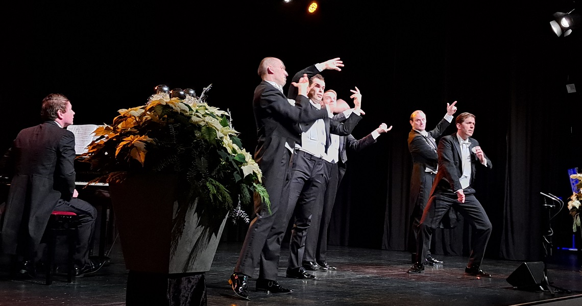 Jubelsturm für „echte“ Doubles der Harmonists Sechs Männer in Frack und Fliege führen mit expressiver Gestik eine komödiantische Bühnenszene auf, während ein Pianist links am Flügel begleitet.