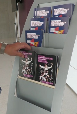 Ein Prospektständer mit Broschüren zur Stadthalle Süßen und mehreren Exemplaren des Programmhefts „Ein rätselhafter Schimmer“. Eine Hand greift nach einem Heft.