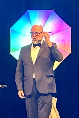 Mann im Anzug und Fliege steht vor einem bunt beleuchteten Schirm auf der Bühne.