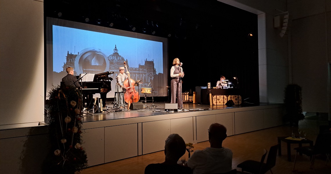 EIN RÄTSELHAFTER SCHIMMER Bühnenaufnahme in der Kulturhalle Süßen: Sängerin mit Mikrofon, begleitet von Klavier, Kontrabass und weiteren Musikern, vor einer Projektion historischer Gebäude. Foto: Katharina Weissenborn.
