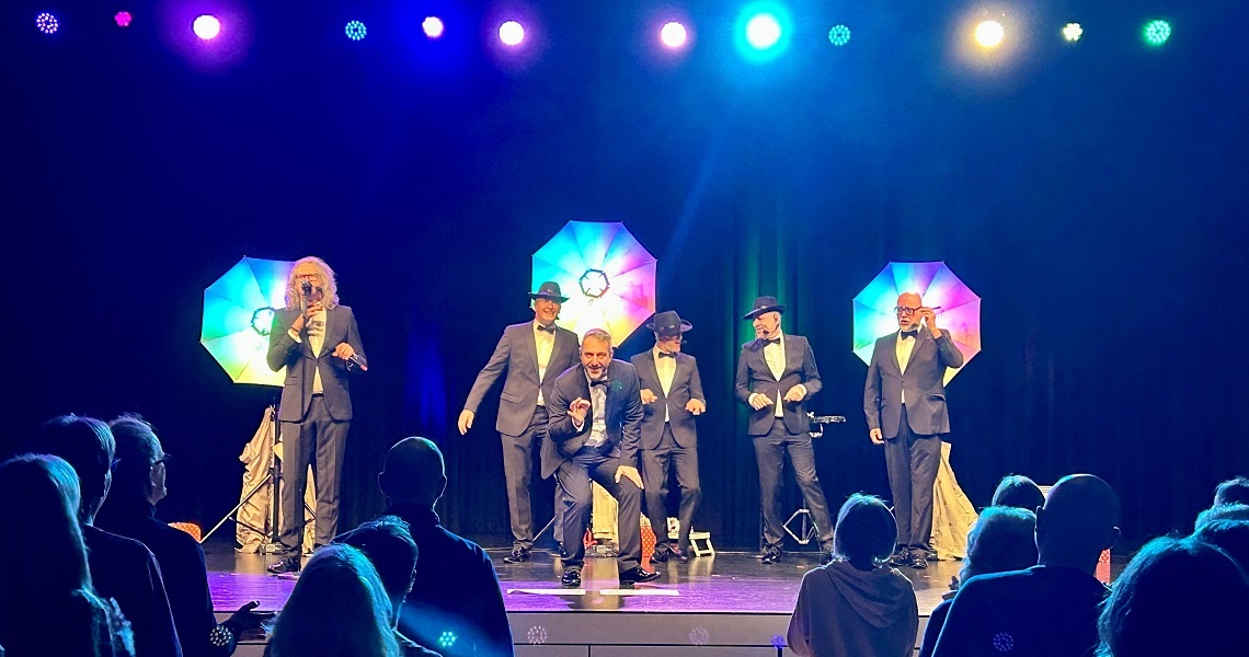 A Cappella Comedy GOLDSINGER Fünf Männer im Anzug und Hut performen auf einer Bühne vor einem begeisterten Publikum. Hinter ihnen leuchten drei farbige Bühnenschirme in Regenbogenfarben.
