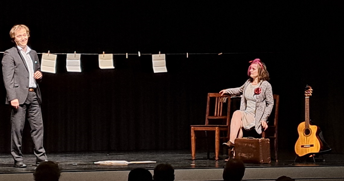 Weiberheld – Mit Tucholsky im Bett Zwei Schauspieler auf einer Theaterbühne: Ein Mann im Anzug steht links, eine Frau in einem grauen Kostüm mit pinker Schleife sitzt rechts auf einem Koffer. Zwischen ihnen ist eine Wäscheleine mit beschrifteten Blättern gespannt. Eine Akustikgitarre lehnt am rechten Bühnenrand. Die Szene stammt aus einem Bühnenstück über Kurt Tucholsky.