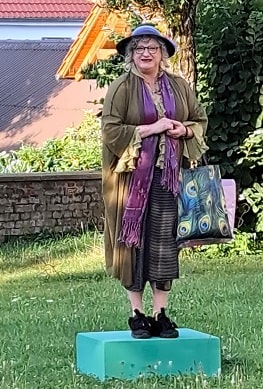 Eine Schauspielerin des Ensembles Dein Theater steht beim poetischen Stadtspaziergang in Süßen auf einem grünen Podest und trägt Hut, Schal und eine farbenfrohe Tasche.
