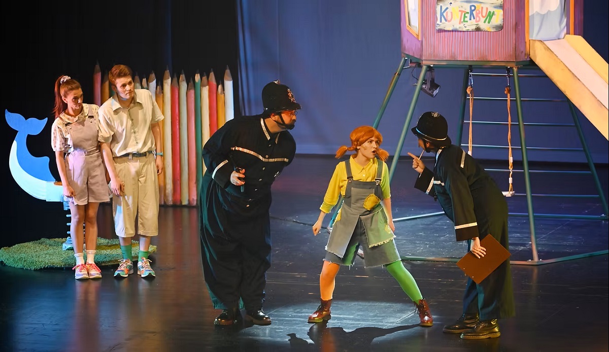 Szene aus dem Familienmusical Pippi Langstrumpf: Ein starkes Mädchen mit roten Zöpfen widerspricht zwei Polizisten auf der Bühne, während zwei Kinder gespannt zuschauen.