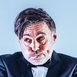 Lars Kienle Mann im dunklen Anzug mit Fliege, humorvoller Gesichtsausdruck, leicht nach unten blickend, vor einfarbigem Hintergrund.