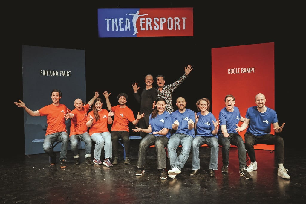 Kulturhalle Süßen präsentiert: THEATERSPORT Zwei Improvisationstheater-Teams in roten und blauen Shirts sitzen jubelnd auf einer Bühne unter dem Schriftzug „Theatersport“, hinter ihnen stehen farblich passende Tafeln mit den Teamnamen „Fortuna Faust“ und „Coole Rampe“.