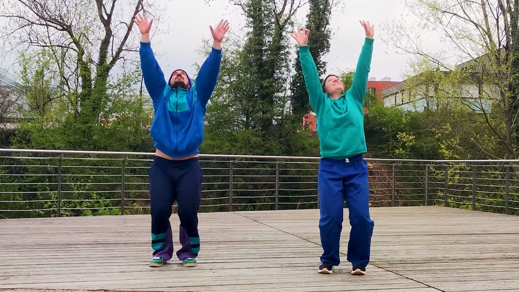Zwei Personen führen im Freien eine Tanzbewegung mit erhobenen Armen als Teil einer Flashmob-Choreografie aus.