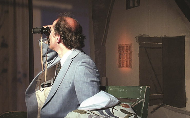 Ein Mann sitzt auf einer Bühne und blickt durch ein Fernglas in die Ferne. Nachmittagstheater: Dositza, bloß so dositza