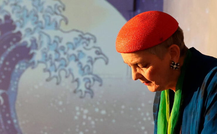Eine Frau mit roter Kopfbedeckung steht im Bühnenlicht vor einer projizierten Welle. Nachmittagstheater: Alles fließt