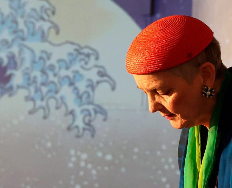 Eine Frau mit roter Kopfbedeckung steht im Bühnenlicht vor einer projizierten Welle. Nachmittagstheater: Alles fließt
