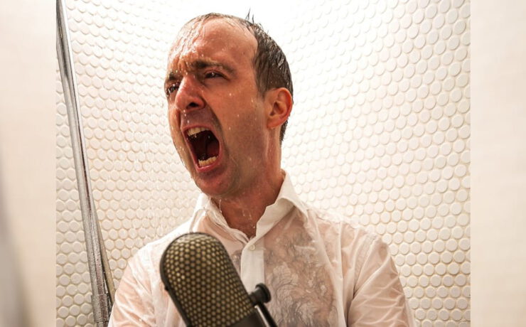 Patrick Bopp singt aus voller Kehle in ein Mikrofon, während Wasser auf ihn herabprasselt – passend zum Mitsinge-Konzert „Aus voller Kehle für die Seele“.