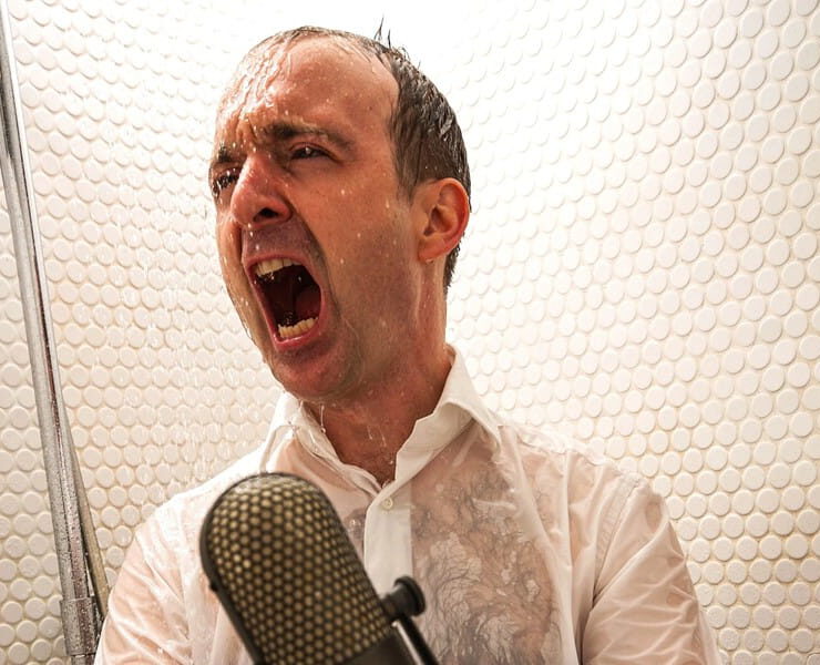 Patrick Bopp singt aus voller Kehle in ein Mikrofon, während Wasser auf ihn herabprasselt – passend zum Mitsinge-Konzert „Aus voller Kehle für die Seele“.