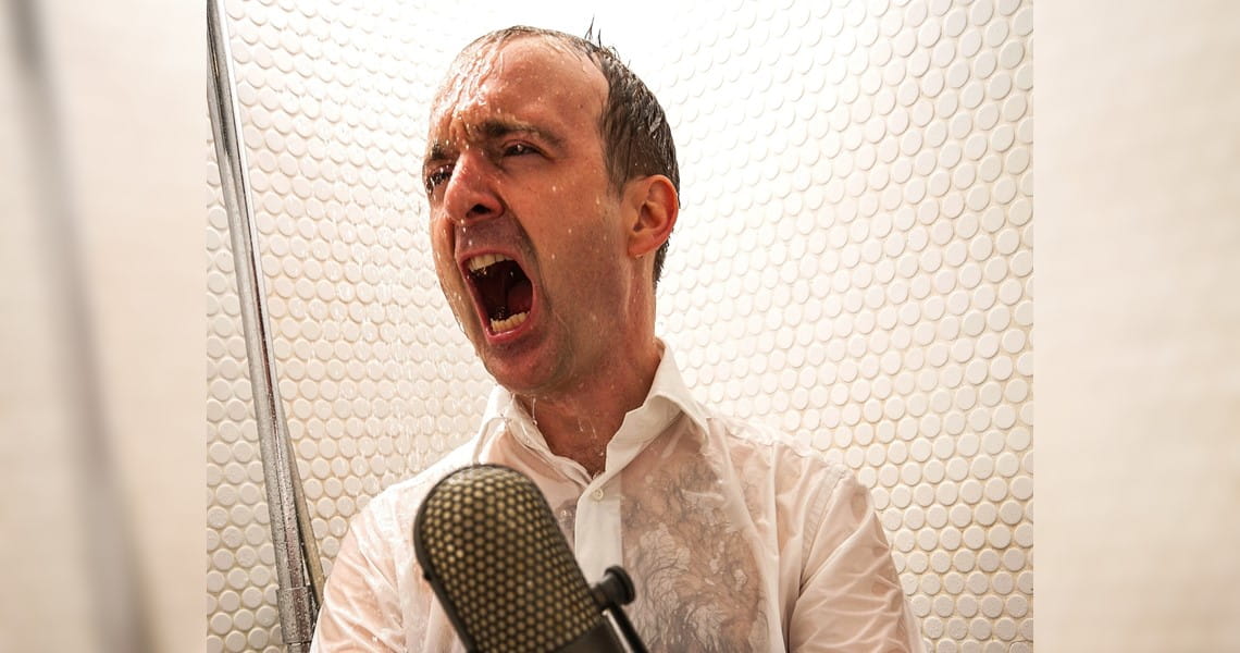 Mitsinge-Konzert zur Sonnenwende | AUS VOLLER KEHLE FÜR DIE SEELE Patrick Bopp singt aus voller Kehle in ein Mikrofon, während Wasser auf ihn herabprasselt – passend zum Mitsinge-Konzert „Aus voller Kehle für die Seele“.