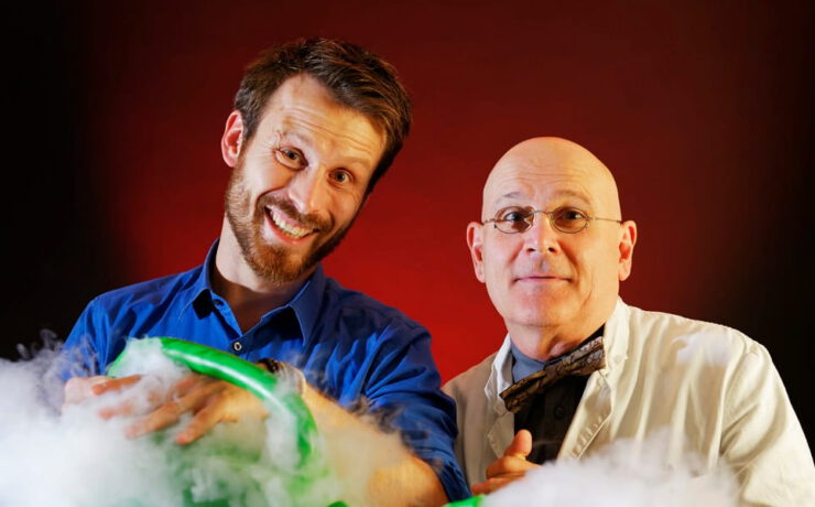 Die Physikanten stehen vor rot-schwarzem Hintergrund zwischen aufsteigendem Nebel und einem grün leuchtenden Experiment – passend zur Wissensshow „Best of Weihnachten“.