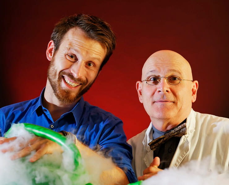 Die Physikanten stehen vor rot-schwarzem Hintergrund zwischen aufsteigendem Nebel und einem grün leuchtenden Experiment – passend zur Wissensshow „Best of Weihnachten“.