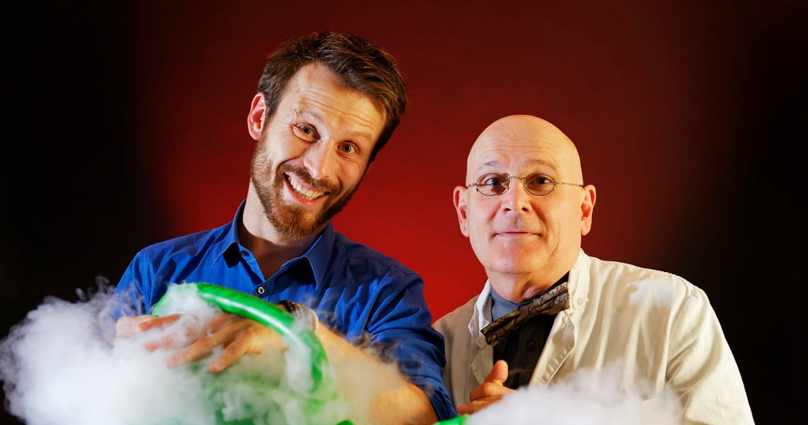 Die interaktive Wissensshow | BEST OF WEIHNACHTEN Die Physikanten stehen vor rot-schwarzem Hintergrund zwischen aufsteigendem Nebel und einem grün leuchtenden Experiment – passend zur Wissensshow „Best of Weihnachten“.