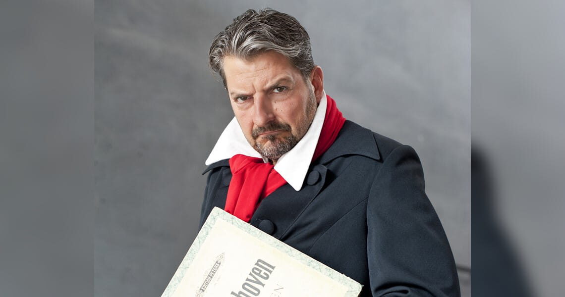 Felix Reuter posiert in Beethoven-Kostüm mit rotem Schal und Notenblatt vor grauem Hintergrund – passend zur Musikcomedy „Der verflixte Beethoven“.