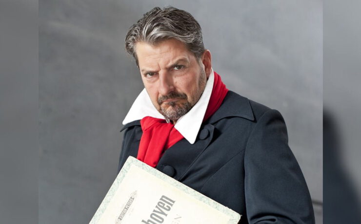 Felix Reuter posiert in Beethoven-Kostüm mit rotem Schal und Notenblatt vor grauem Hintergrund – passend zur Musikcomedy „Der verflixte Beethoven“.
