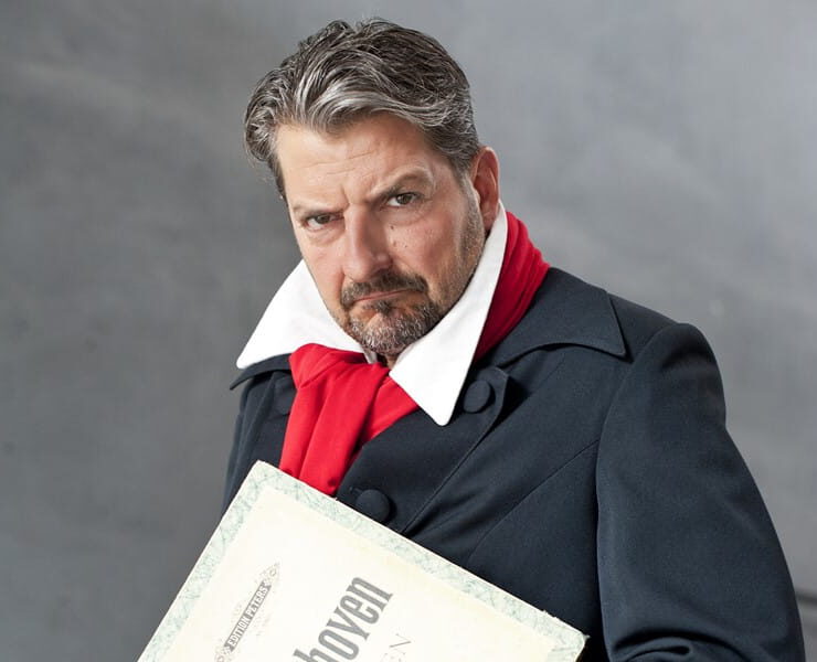 Felix Reuter posiert in Beethoven-Kostüm mit rotem Schal und Notenblatt vor grauem Hintergrund – passend zur Musikcomedy „Der verflixte Beethoven“.