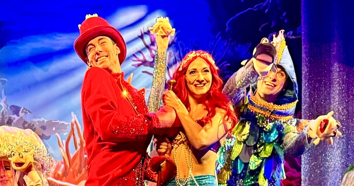 Szene aus dem Familienmusical „Die kleine Meerjungfrau“ mit farbenfrohen Kostümen, Unterwasser-Bühnenbild und Figuren wie Meerjungfrau, Krabbe und Fisch.