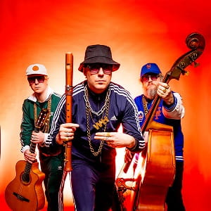 Wildes Holz – Blockflöte, Gitarre und Kontrabass mit Haltung Die Band Wildes Holz posiert vor orangefarbenem Hintergrund. Die drei Musiker tragen lässige Kleidung und Hüte; im Vordergrund hält Tobias Reisige eine Blockflöte, links sitzt Johannes Behr mit Gitarre, rechts steht Markus Conrads mit Kontrabass.