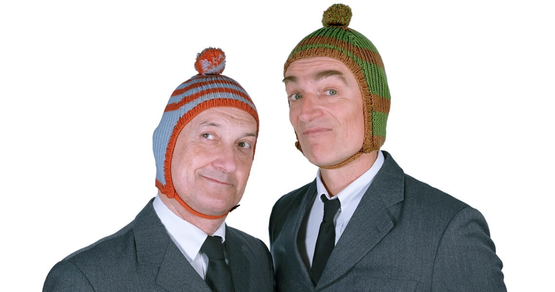 Die beiden Künstler von Ulan & Bator posieren in grauen Anzügen mit bunten Strickmützen und schauen verschmitzt in die Kamera.