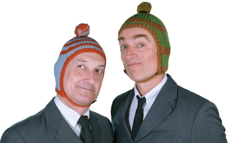 Die beiden Künstler von Ulan & Bator posieren in grauen Anzügen mit bunten Strickmützen und schauen verschmitzt in die Kamera.